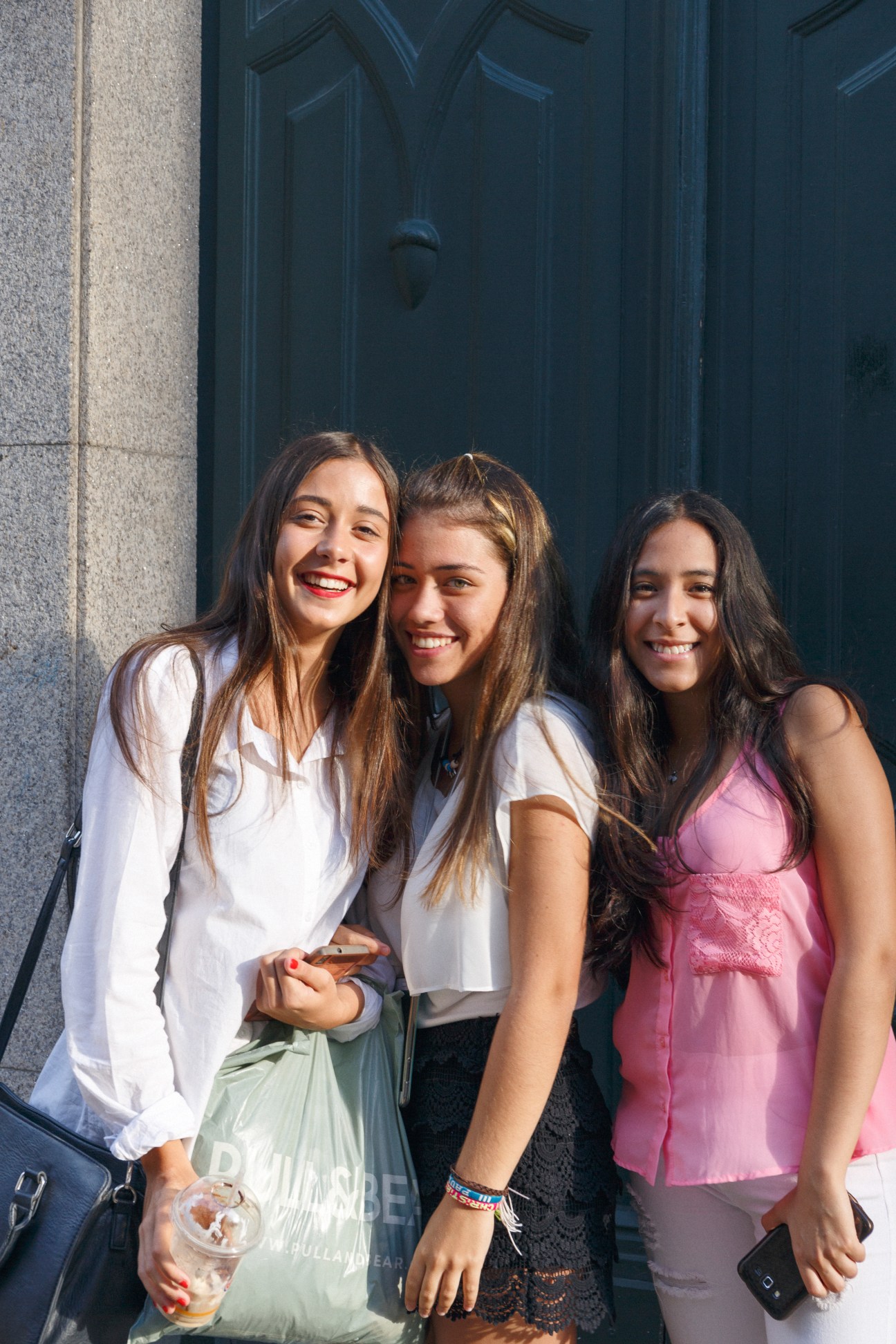 Three young women smiling at camera on a city street with dark blue door behind them.