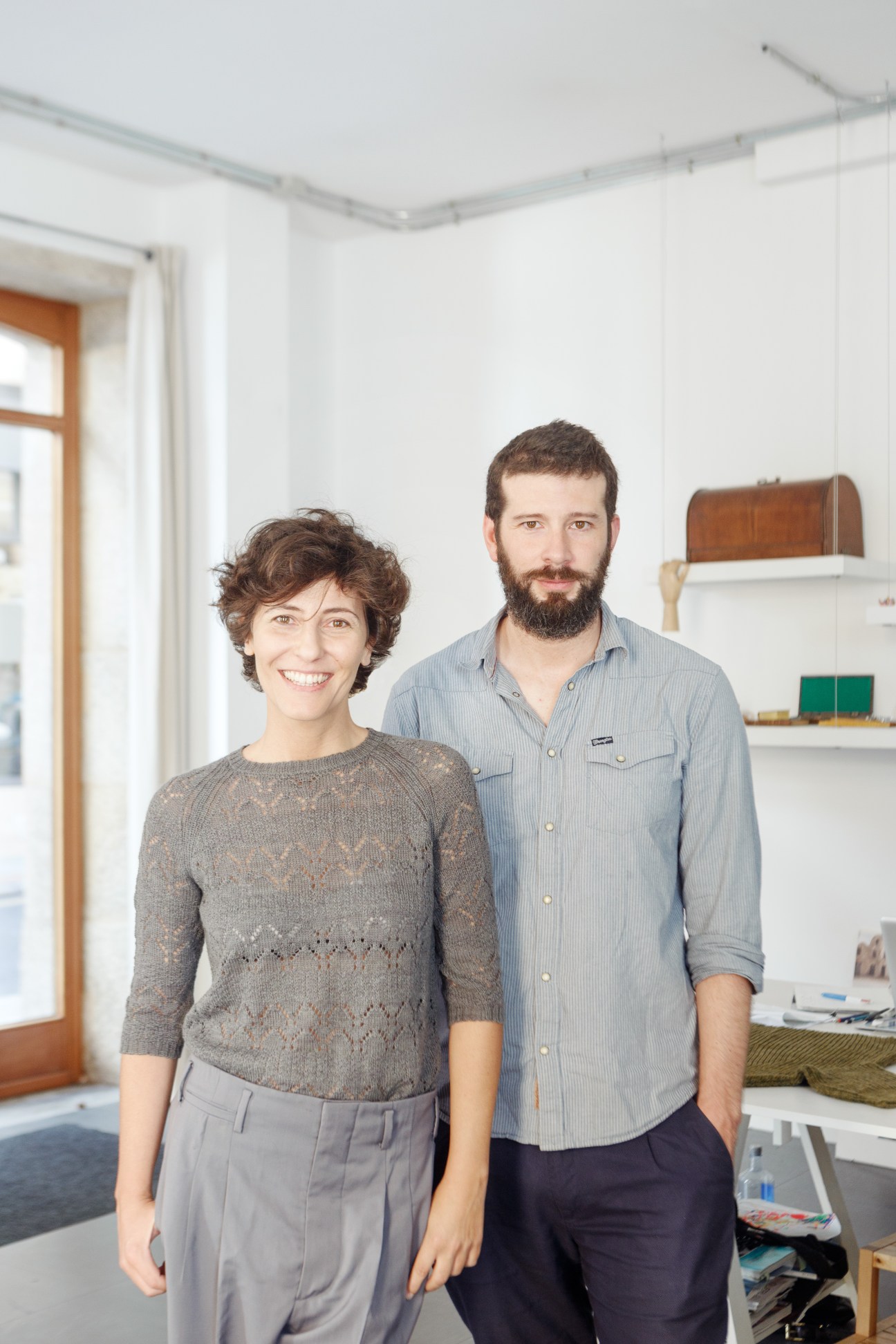 Knitbrary founders Yolanda Estévez and Pedro Castellanos standing together in a bright studio space with shelving behind them.