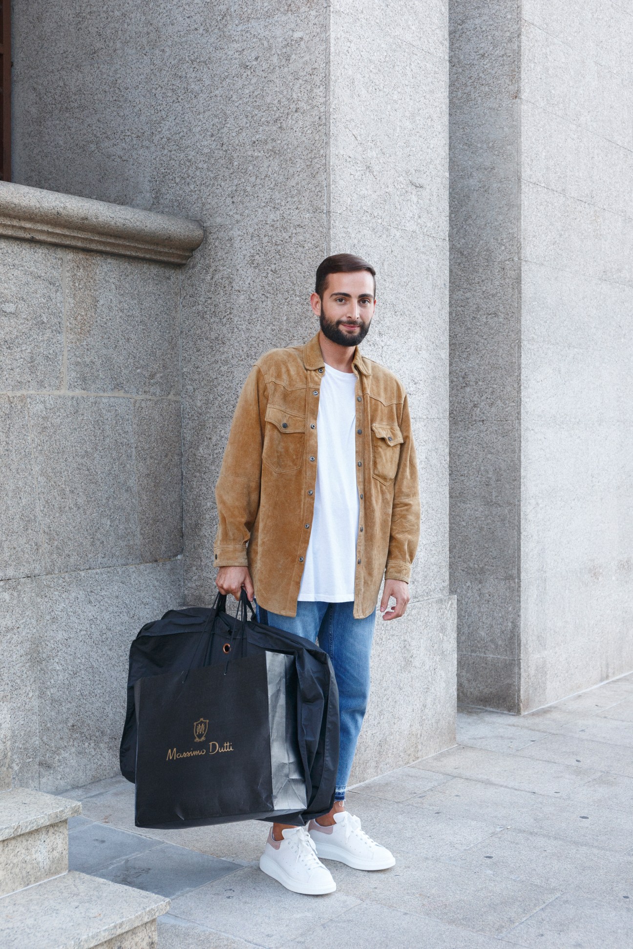 Man in tan suede shirt holding Massimo Dutti bag against stone wall.