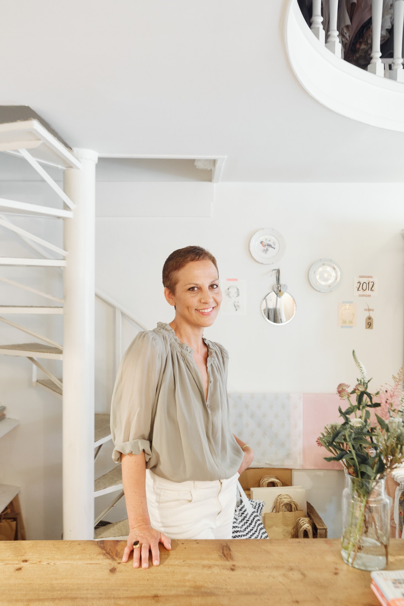 Cristina Pardo, owner of children's boutique Dandelion, smiles while standing at a wooden counter in her minimalist white interior shop.