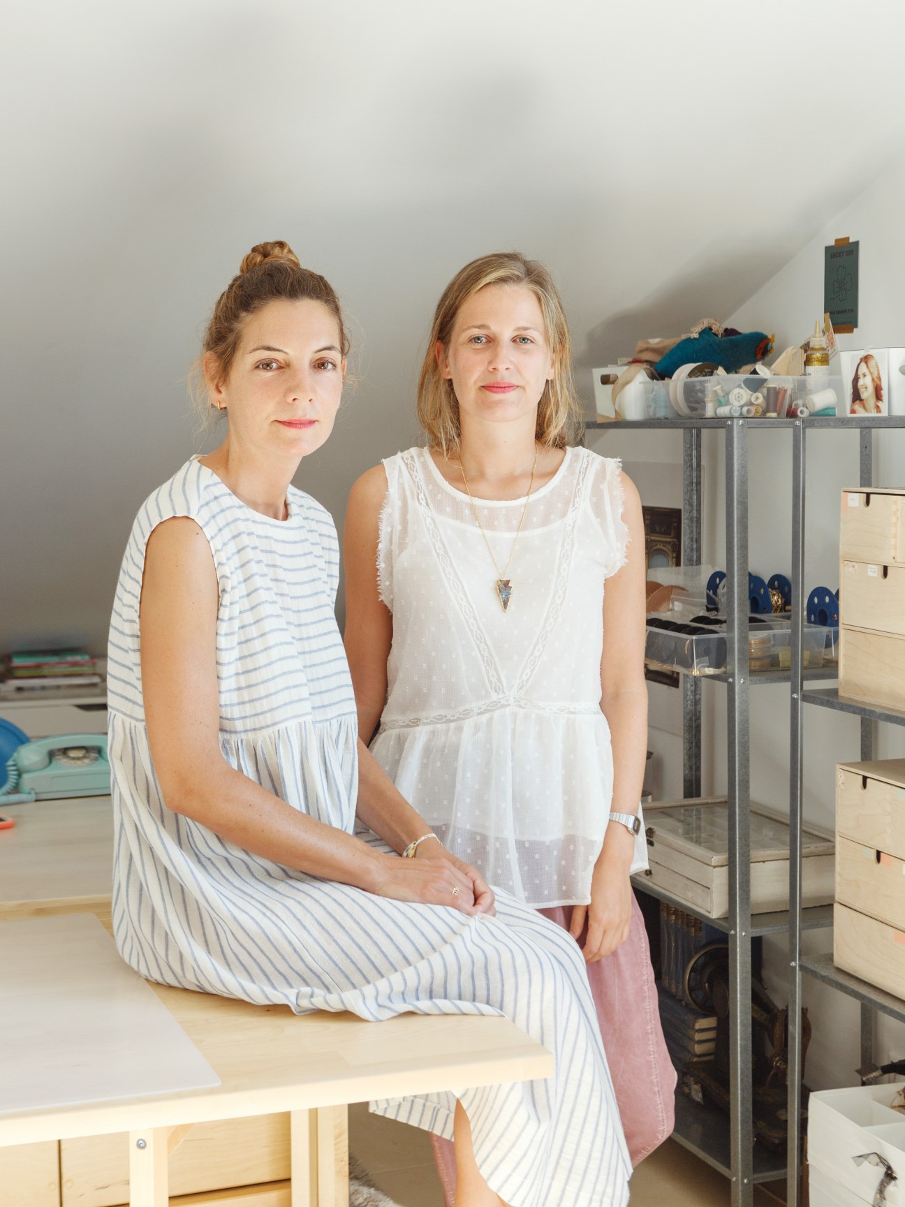 Two women posing in a bright studio space with shelving and design materials behind them.