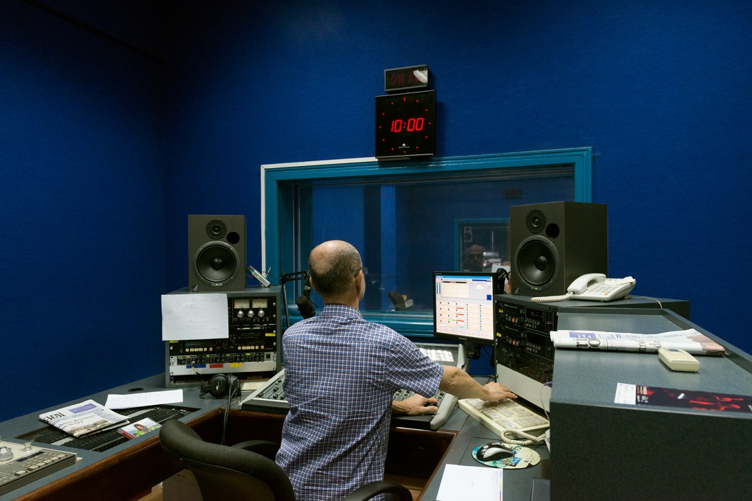 Radio station operator at control console with monitors, speakers, and clock displaying 10:00 in a blue-walled broadcast studio.
