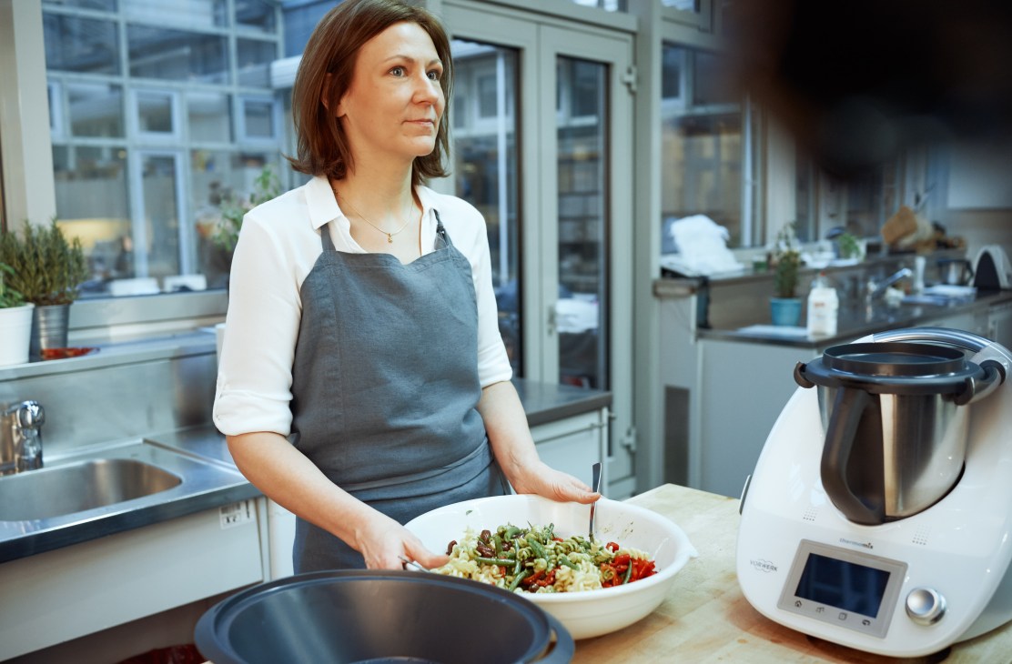 Woman in apron preparing food in modern test kitchen with Thermomix food processor on counter