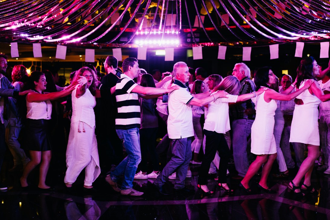 Cruise ship passengers dancing in a conga line on a brightly lit dance floor with decorative lighting overhead.