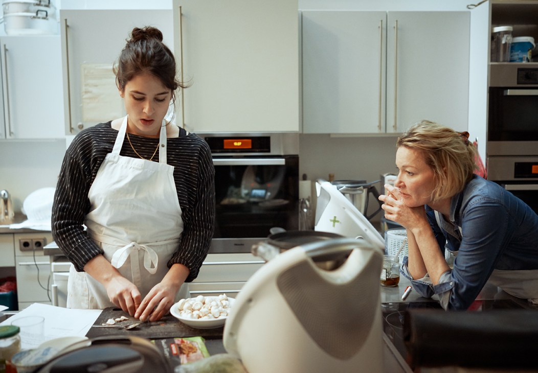 A woman in a white apron chops ingredients in a modern kitchen while another woman watches intently.