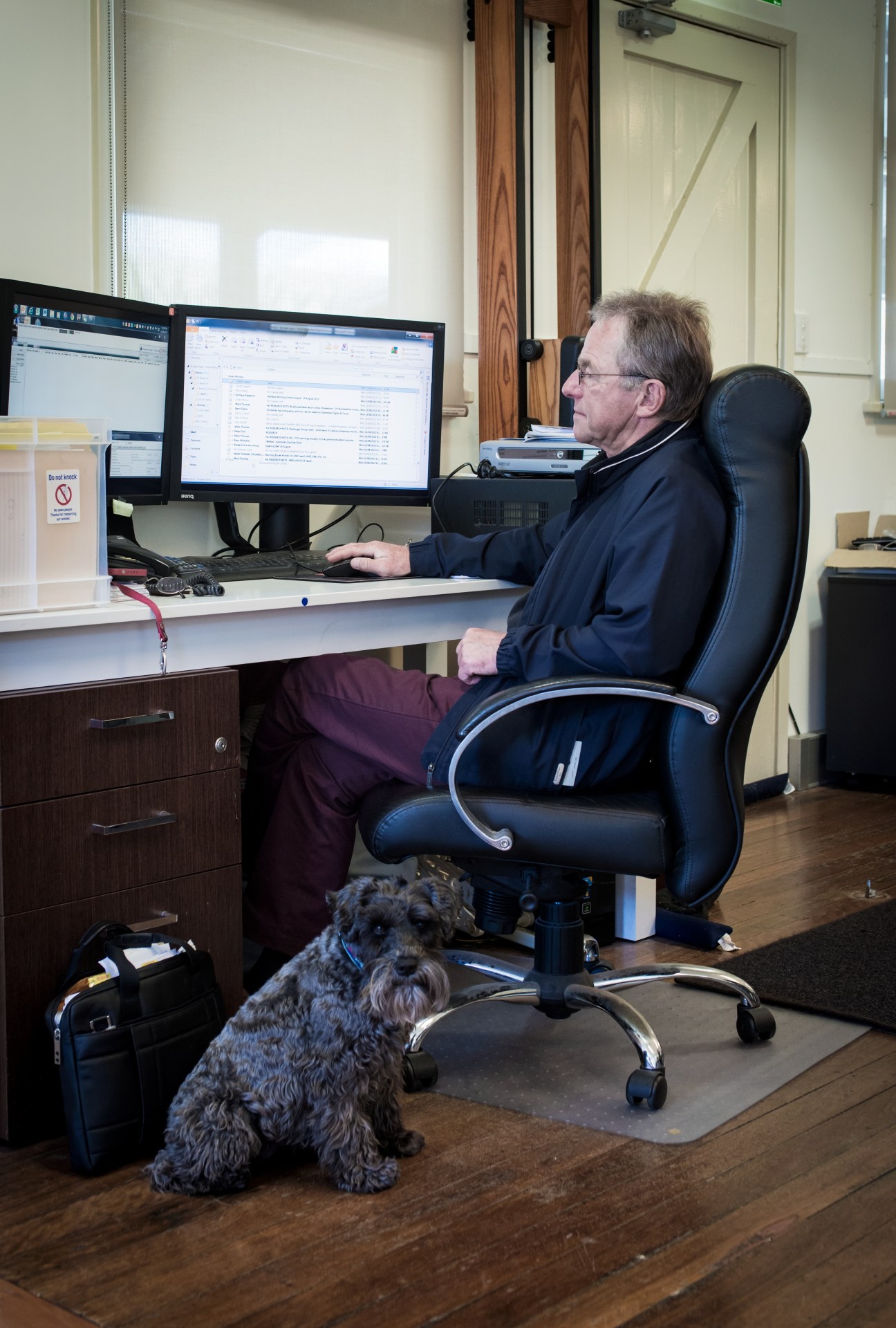 Man working at dual computer monitors at a desk with a dog sitting nearby on the floor.