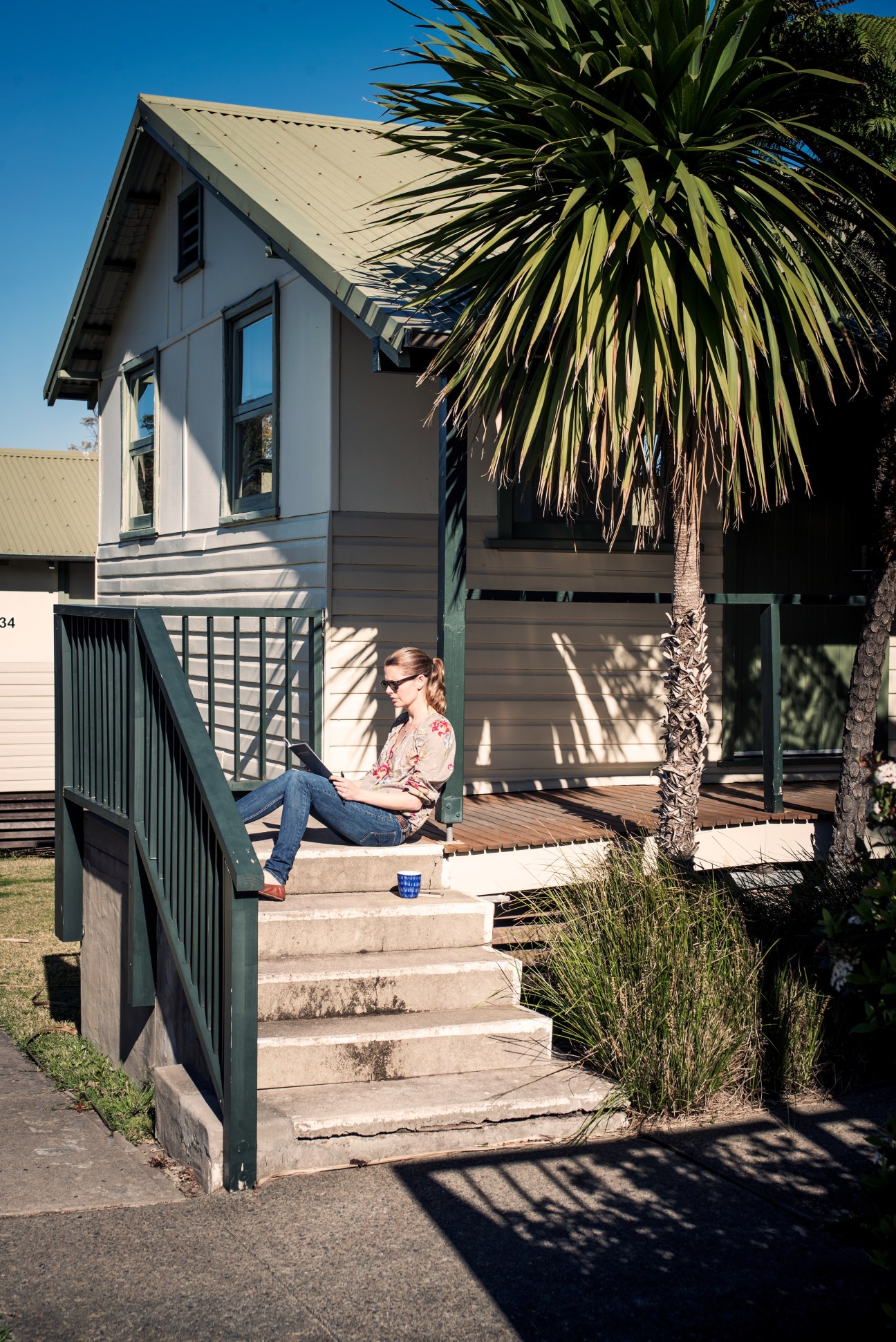 Woman working on laptop outside a white converted hut at Headland Park in Sydney, sitting on steps with palm tree overhead.