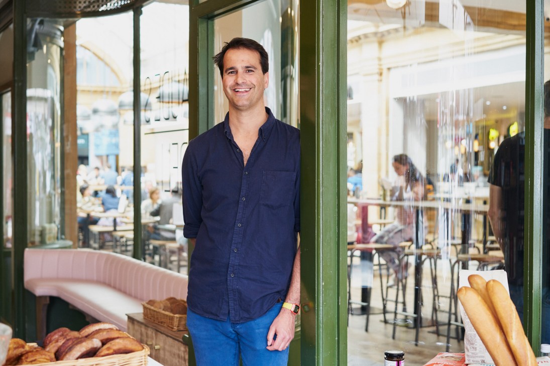 Claude Compton, deli owner, stands smiling in the entrance of Claude's Deli at Market Hall Fulham with fresh bread displayed nearby.