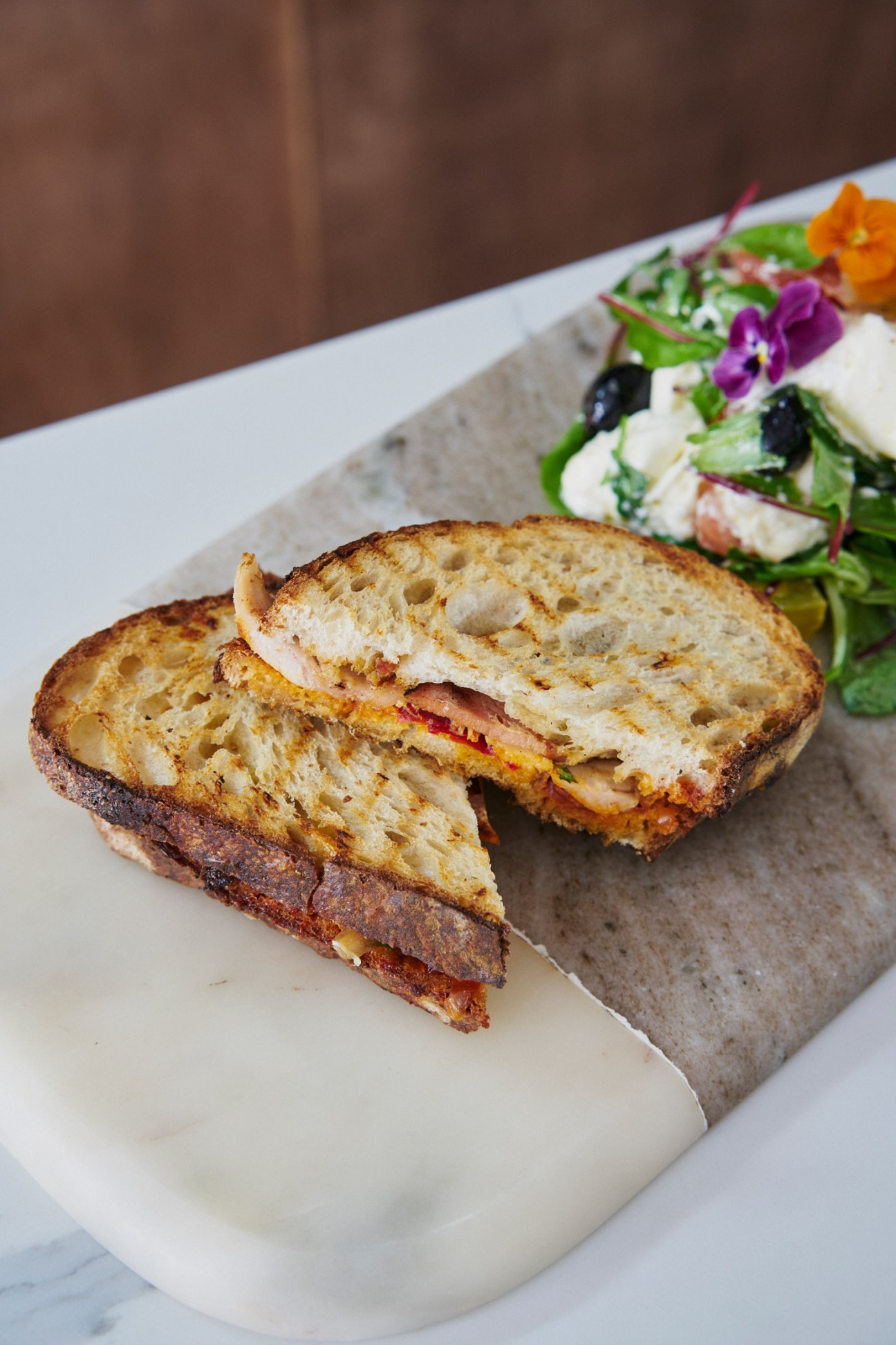 Toasted sandwich with melted cheese and cured meat alongside a salad with edible flowers at Claude's Deli.