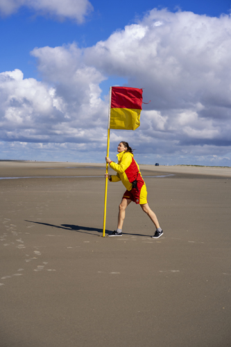 Lifeguard in red and yellow uniform holding rescue flag on windy beach