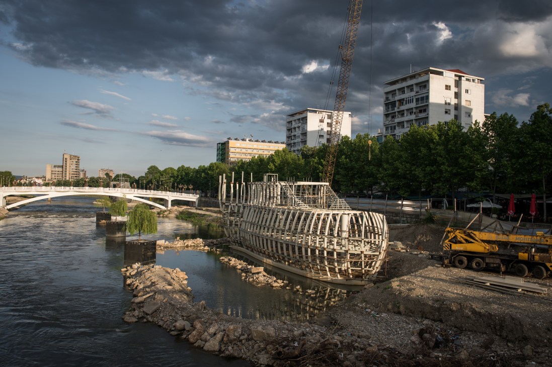 Ship-shaped restaurant structure under construction on the Vardar river in Skopje with modern apartment buildings in the background.