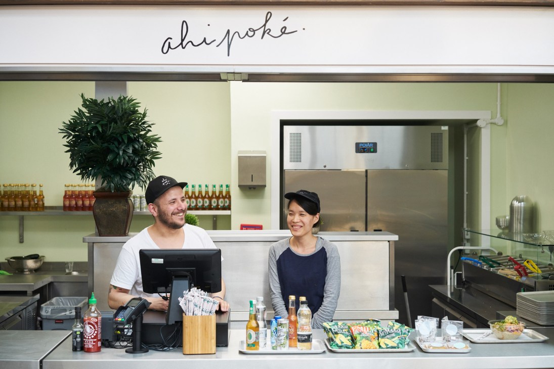 Two staff members smiling behind the counter at Ahi Poké kitchen with light green walls and shelving.