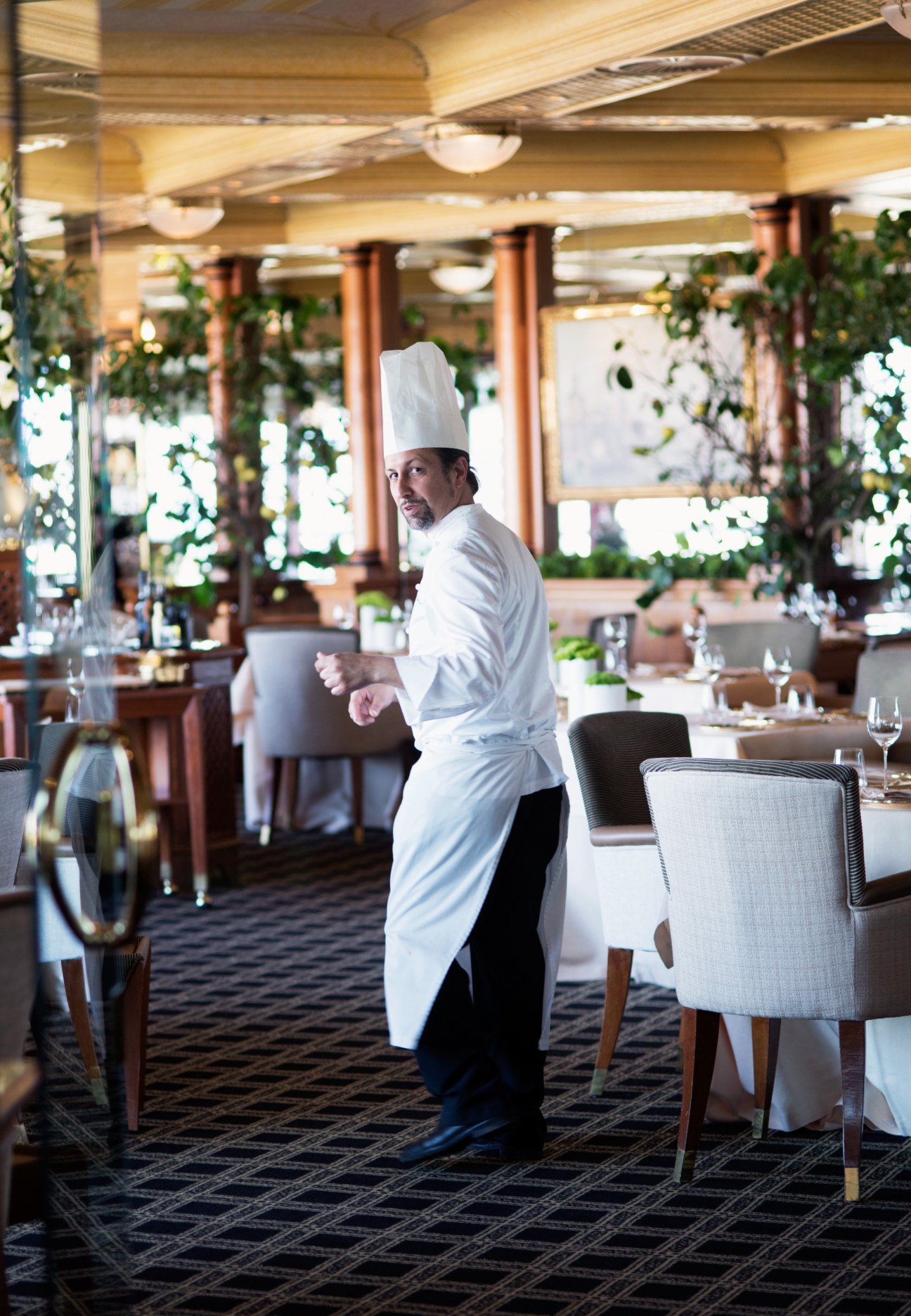 Chef in white chef's coat and hat standing in an elegant dining room at La Pergola restaurant.