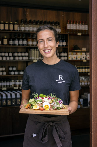 Smiling woman in dark shirt holding wooden board with colorful salad in front of shelved products