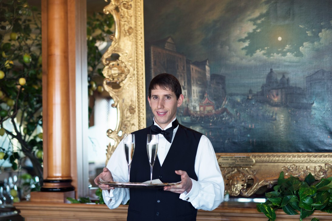 A waiter in formal attire smiles while holding a tray with champagne glasses in an ornate dining room.