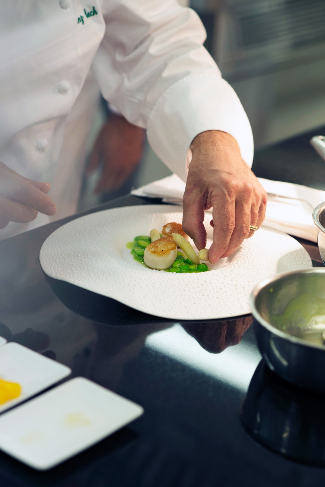 Chef's hand plating a scallop dish with asparagus and peas on white plate.
