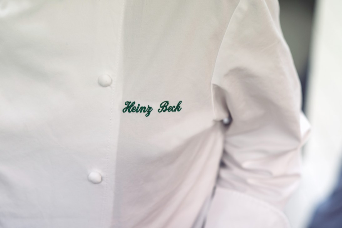 Close-up of a white chef's jacket embroidered with 'Heinz Beck' in green script.