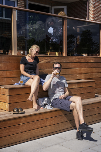 Two people enjoying drinks on wooden steps outside a modern building with glass railings