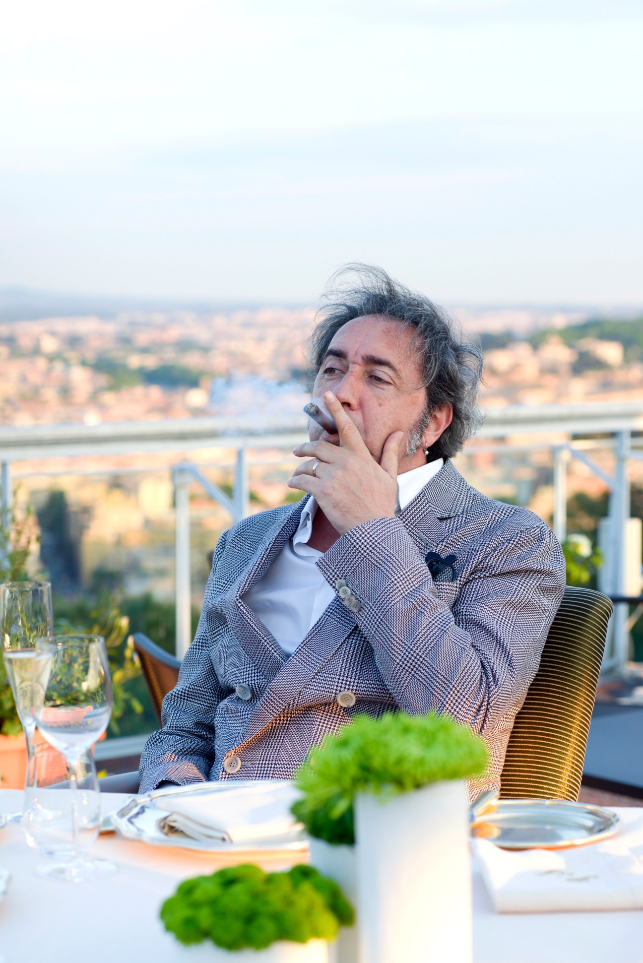 Paolo Sorrentino seated at a dining table overlooking Rome, hand to face in contemplation.