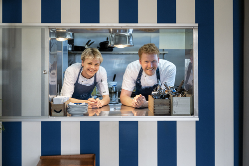 Two chefs in white uniforms and aprons smiling through a kitchen service window