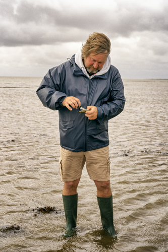 Person in navy jacket and green wellington boots examining something in their hands on sandy beach