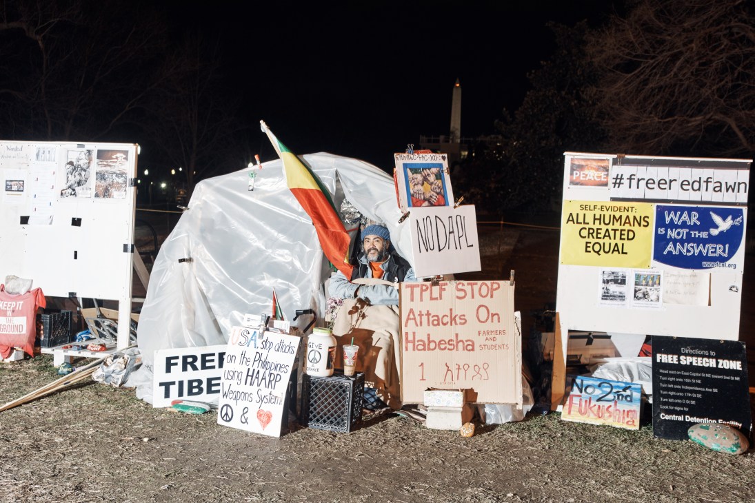 A protestor sitting in a tent encampment near the White House with various signs and flags displayed.