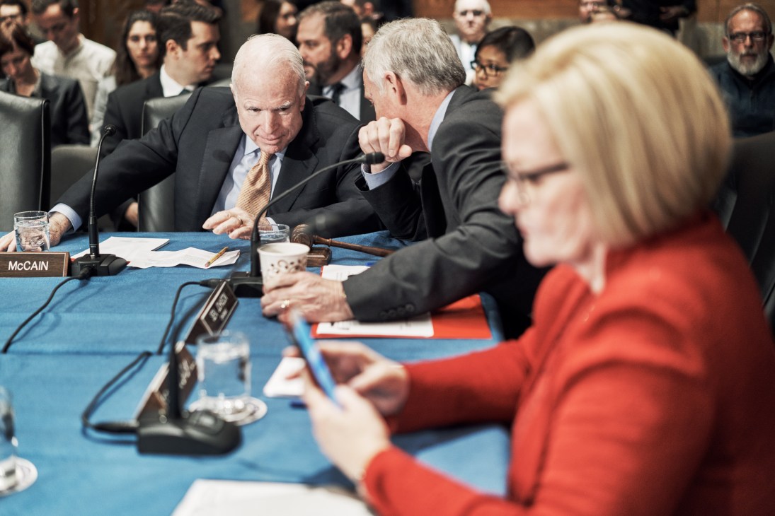 Senator John McCain in conversation at a Senate hearing with other officials seated at a table with microphones.