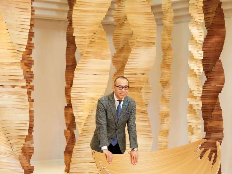 Conde House president Tetsuya Fujita stands beside Jakob Jørgensen's wooden Barca lounge chair in a bright showroom with stacked wood slats.