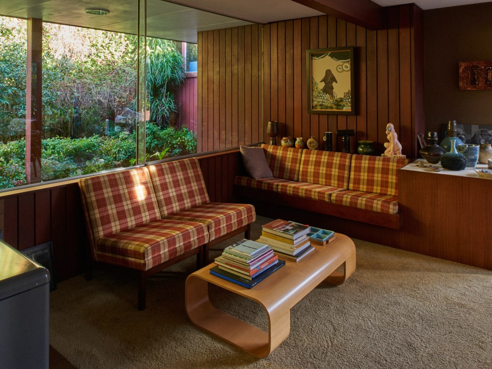 Modernist living room with built-in seating upholstered in red and yellow plaid fabric, wooden walls, and floor-to-ceiling windows overlooking garden.