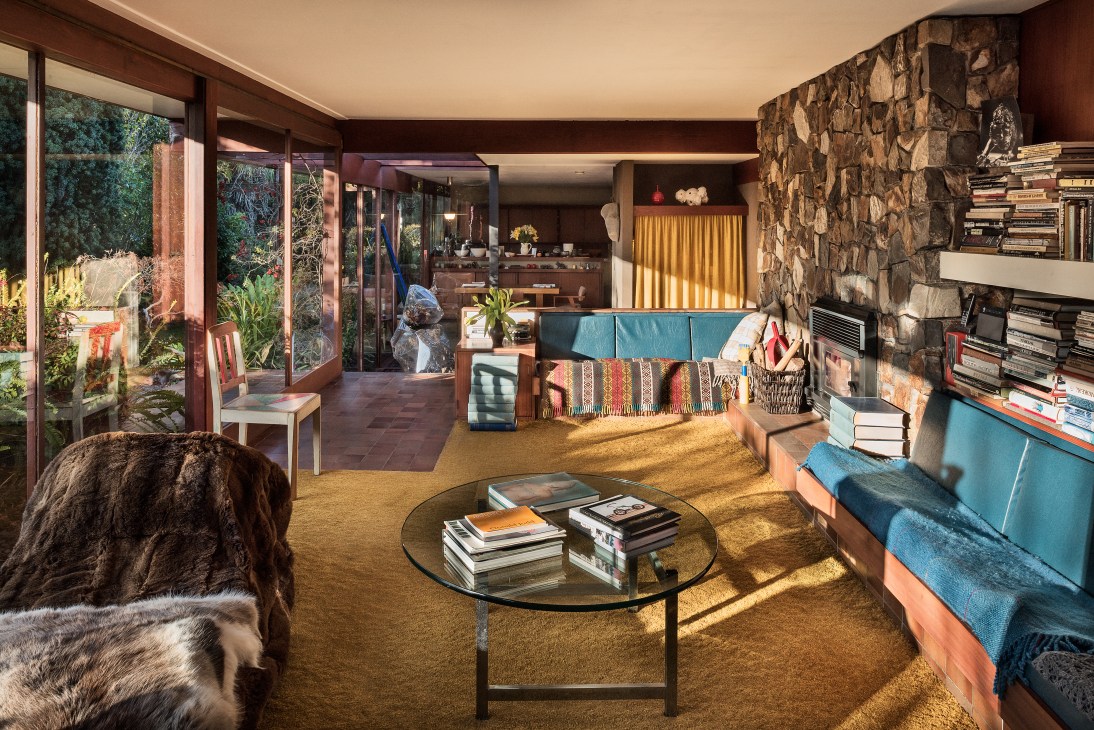 Modernist sitting room with floor-to-ceiling glass doors opening onto garden, stone fireplace, built-in seating, and natural light flooding the space.