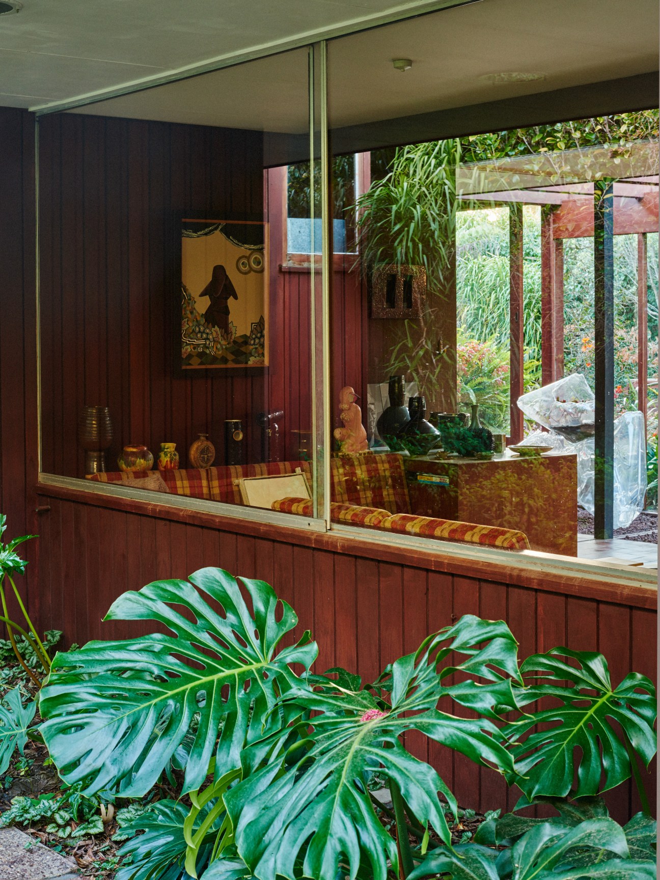 Modernist house exterior with floor-to-ceiling glass windows, redwood cedar paneling, and lush garden plantings in foreground.