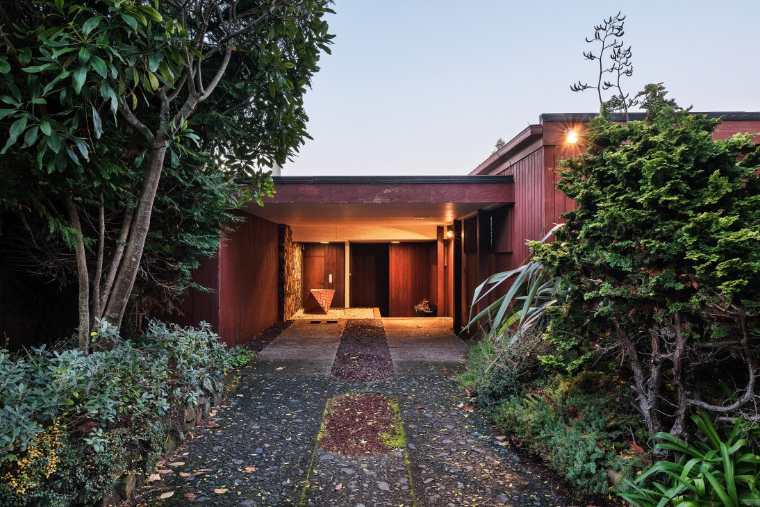 Evening approach to a modernist single-storey home with redwood cedar exterior, surrounded by lush vegetation and illuminated entrance.