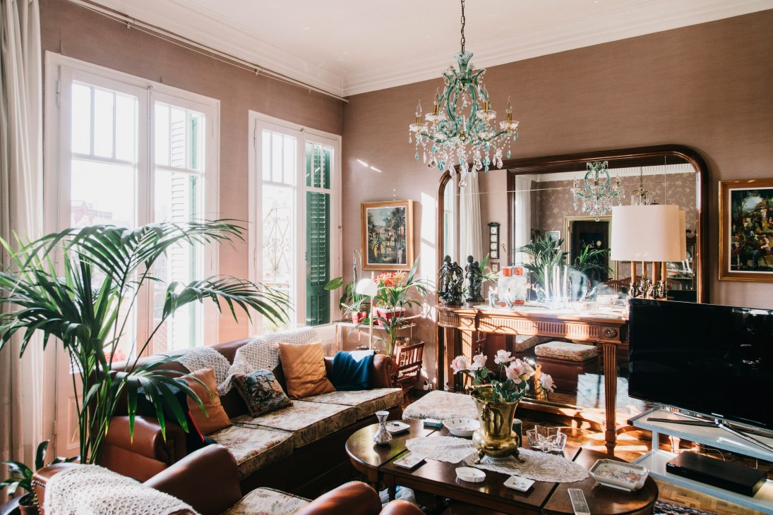 Sunlit living room with terracotta walls, ornate turquoise chandelier, patterned sofa, potted plants, and classical furnishings.