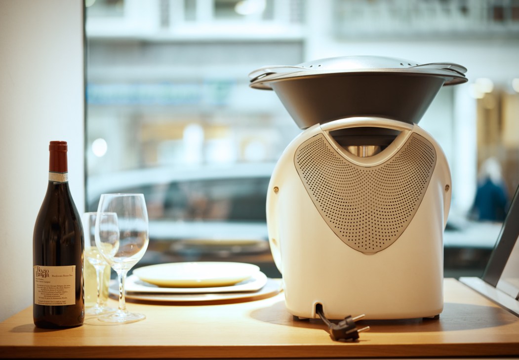 A cream-colored Thermomix food processor with its bowl lid on a dining table, surrounded by wine bottle and glasses.