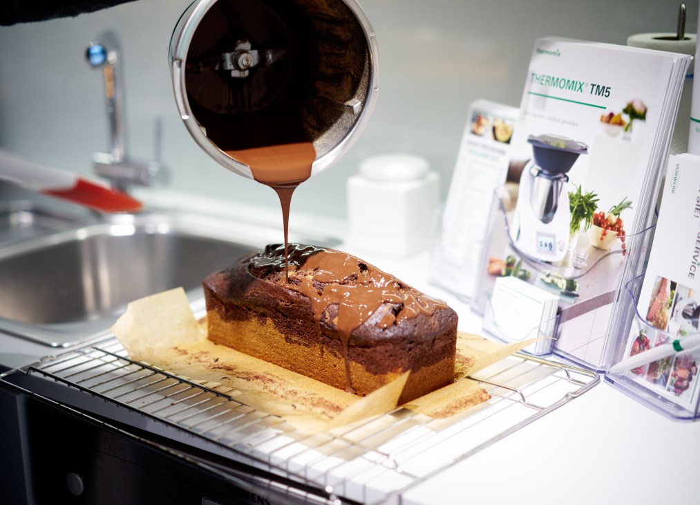 Thermomix TM5 pouring chocolate ganache over a baked cake in a test kitchen.