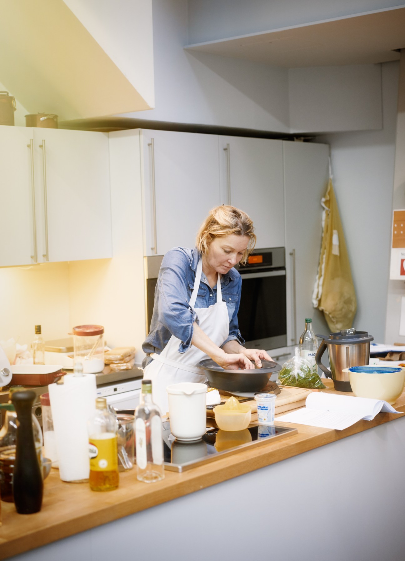 A chef in apron and denim shirt works at a counter in a modern test kitchen, preparing ingredients.
