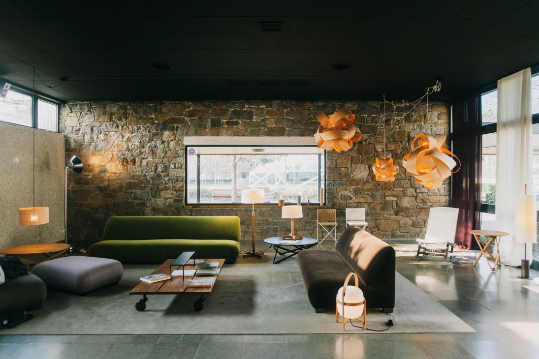 Santa & Cole showroom with green Cadaqués sofas, black armchair, orange Leonardo pendant lamps, and stone wall backdrop.