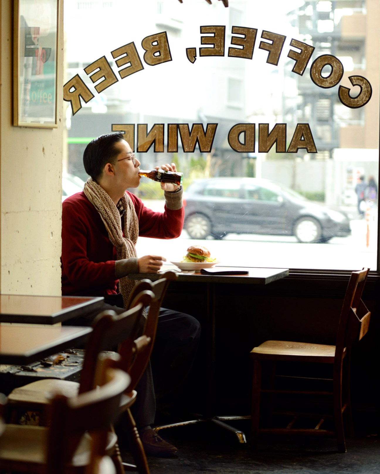 Person in red cardigan and beige scarf sitting at café counter drinking beverage with hamburger plate.