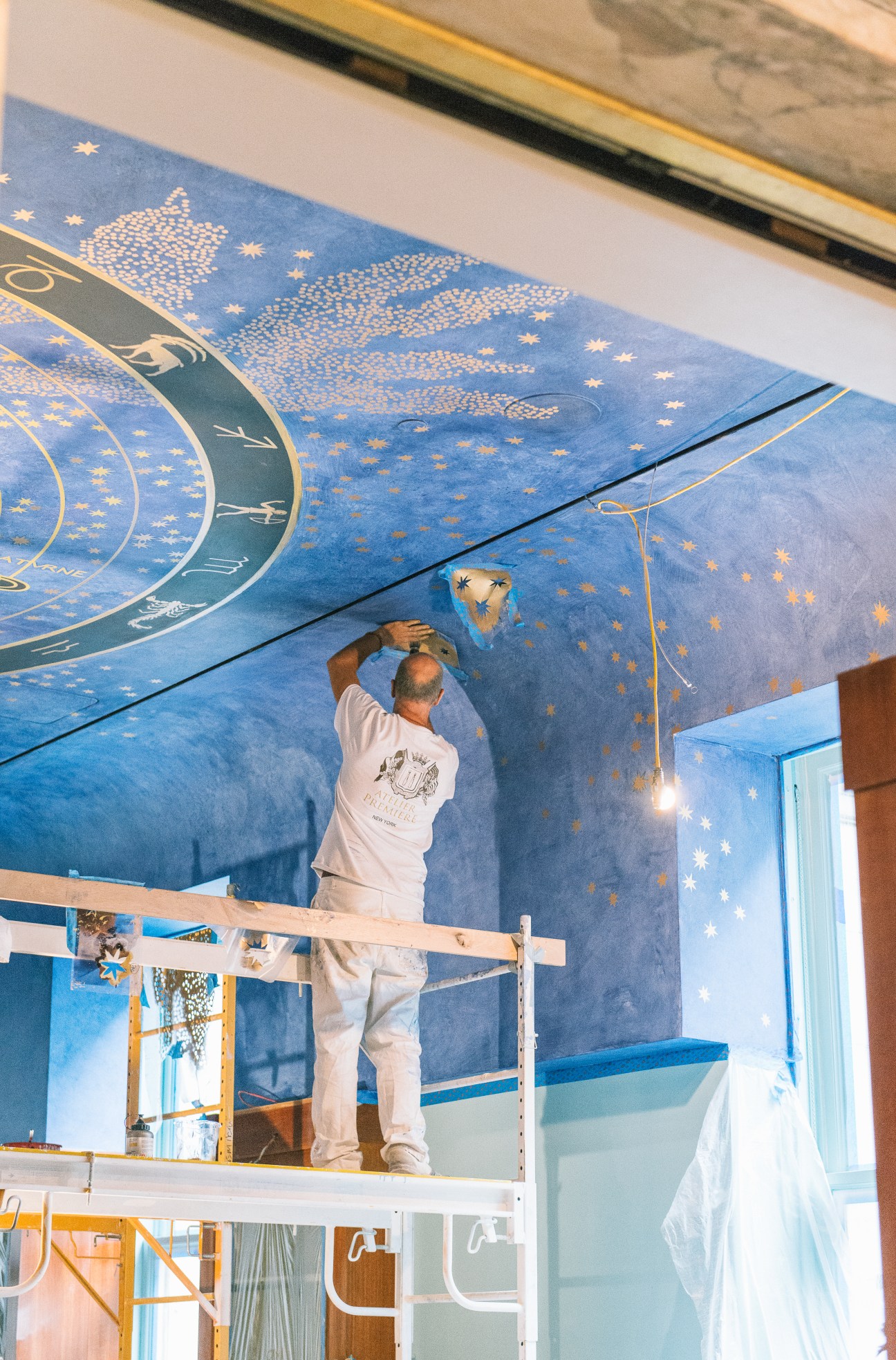 Worker on scaffolding applies finishing touches to a blue and gold fresco ceiling with stars and classical designs.