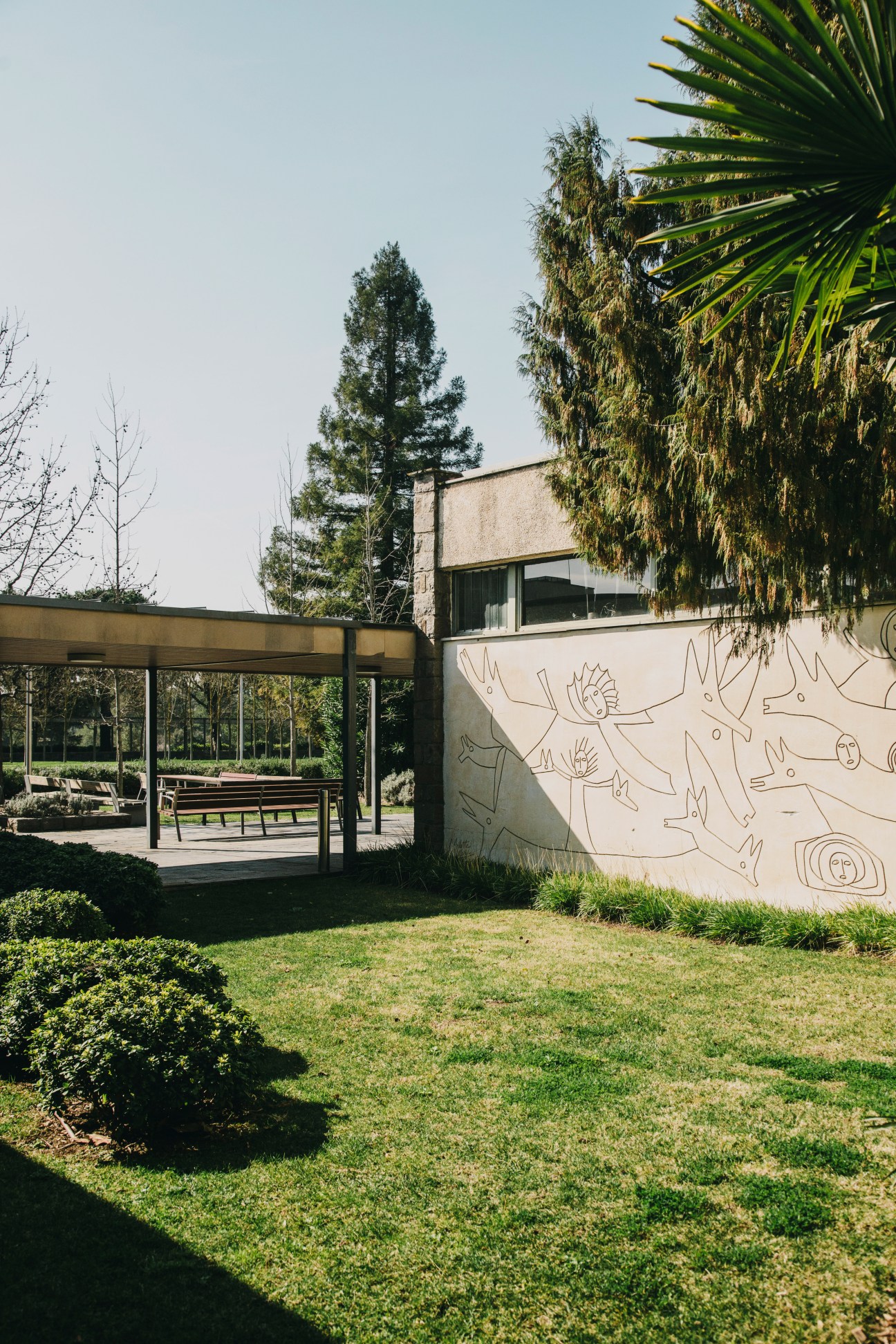Modern building with mural wall art by Arlette Martí, featuring abstract line drawings and ivy-covered architectural elements in a garden setting.