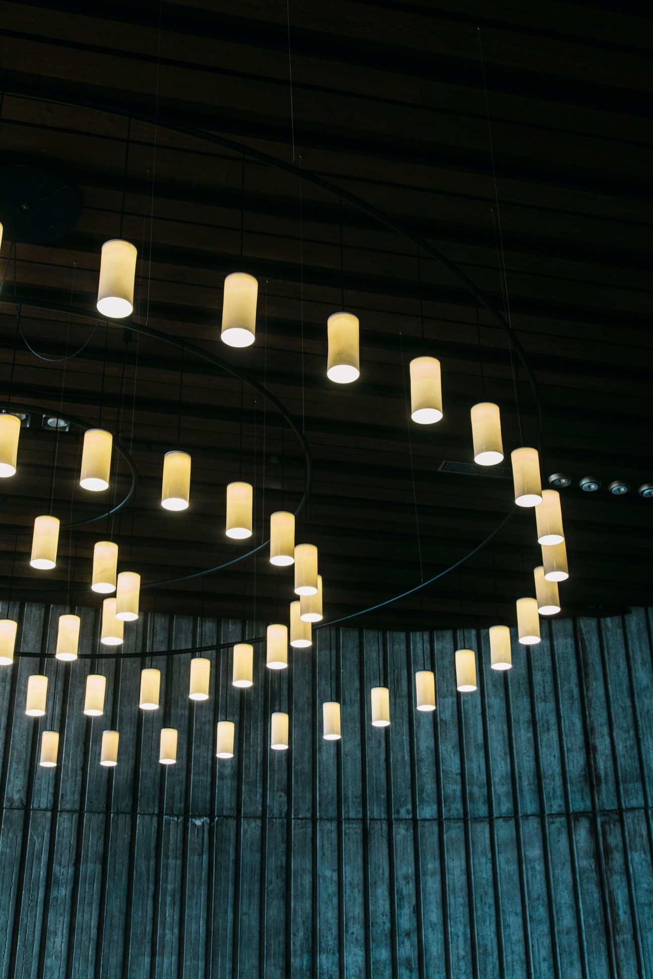 Multiple pendant lamps with cream cylindrical shades hanging from curved wires against dark wooden ceiling.