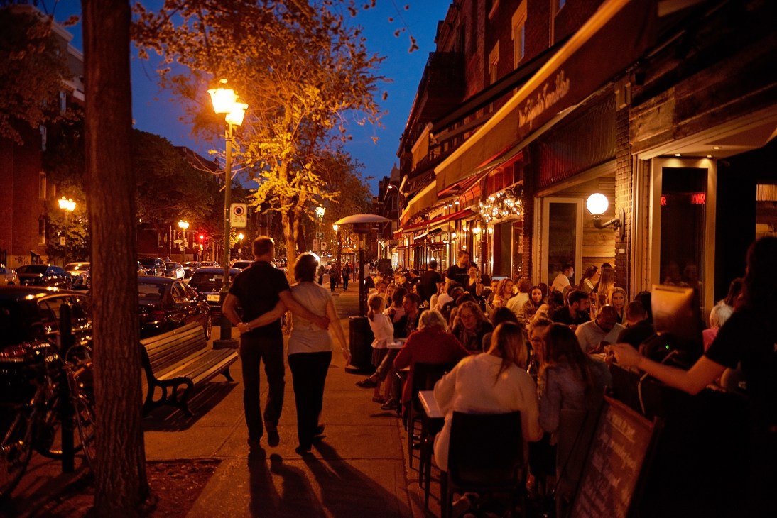 Evening street scene with crowds of people dining and socializing outside illuminated storefronts on Bernard Avenue.