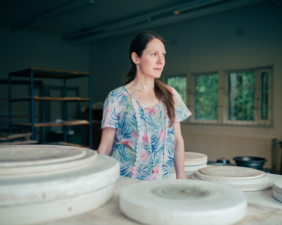 Henna Lamberg stands in a bright studio surrounded by white ceramic pieces and a blue metal shelving unit in the background.