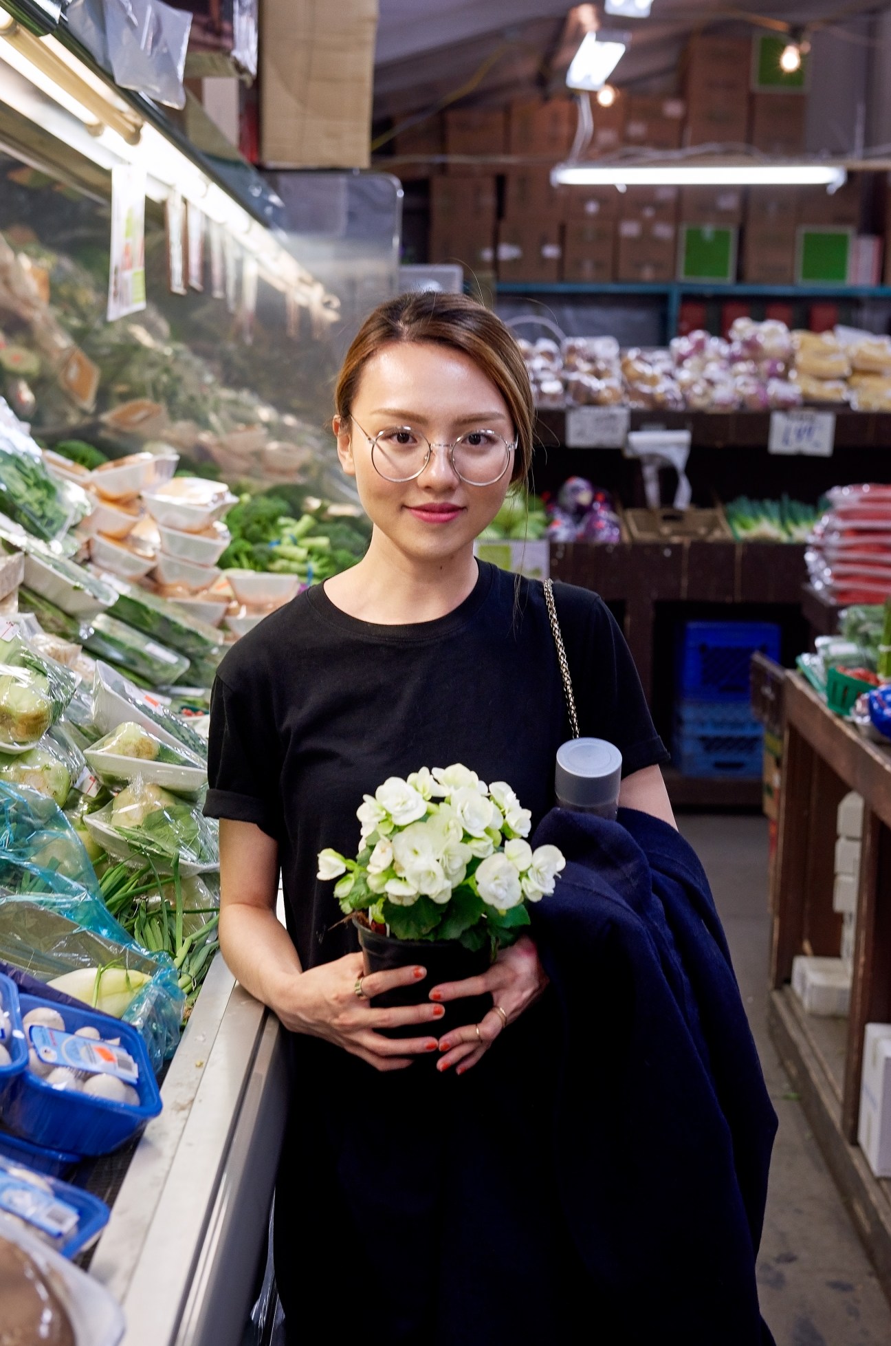 Woman holding white flowers in a late-night market with produce displays and refrigerated cases.