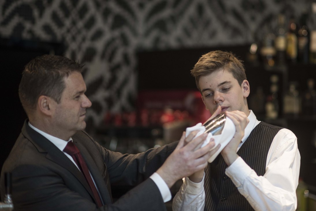 An instructor demonstrates cocktail shaker technique to a young student in formal hospitality attire.