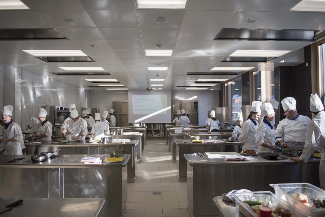 Students in white chef uniforms and hats working at multiple stainless steel kitchen stations during a cooking class.