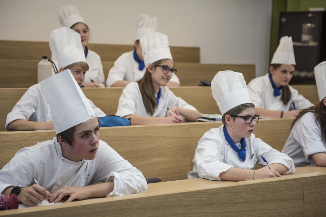Future chefs in white chef's coats and tall white hats sit at wooden desks in a classroom at Brunico Hotel School.