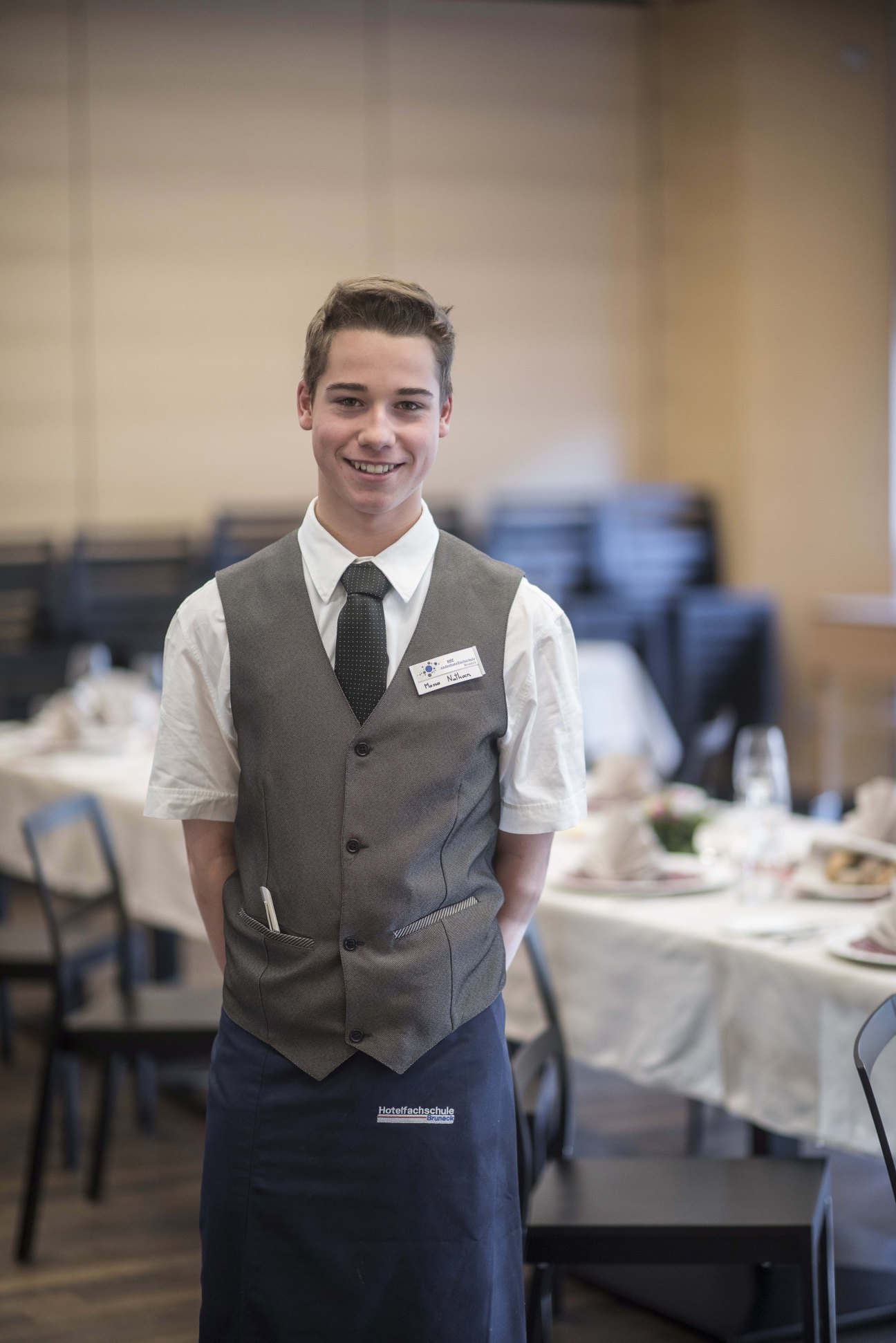 Nathan Massa, a Brunico Hotel School student and future maître d', smiles in formal hospitality uniform in the school's restaurant.
