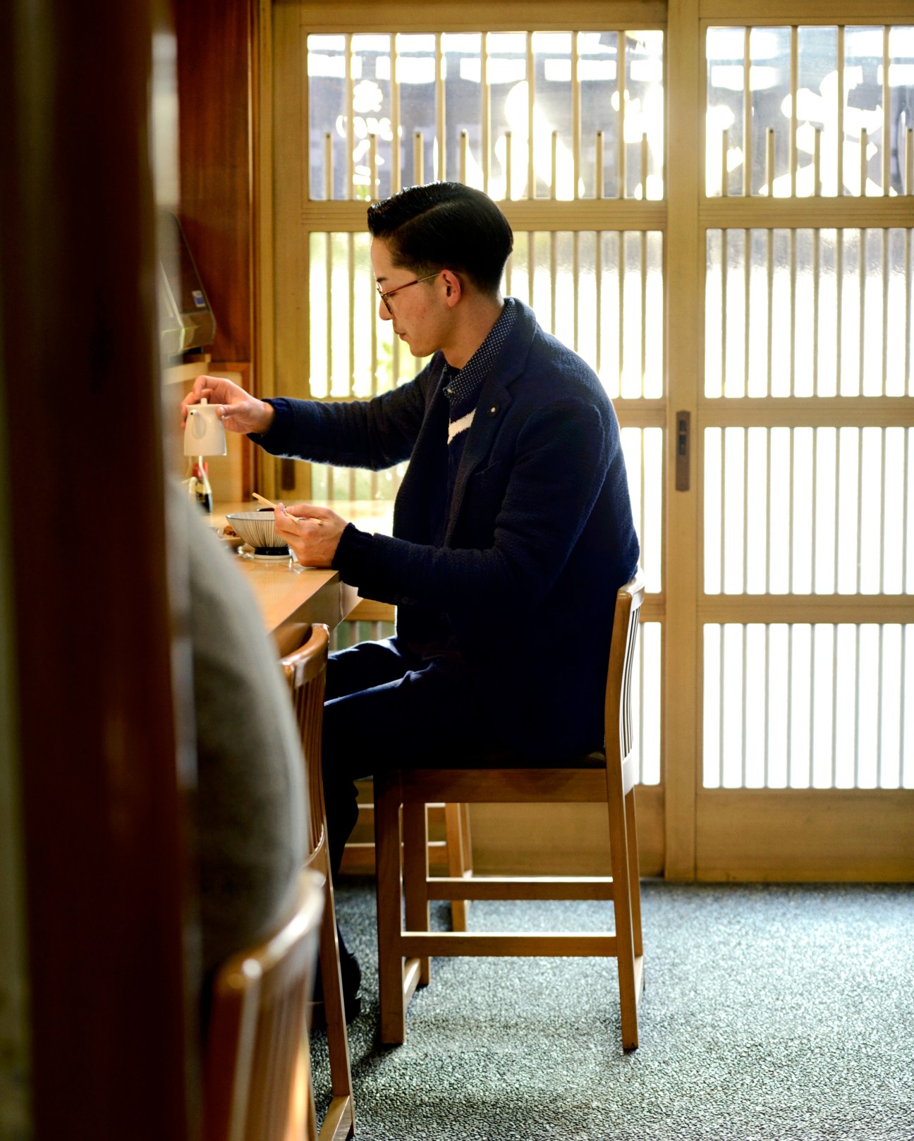 Man in navy cardigan and glasses sits at counter eating at Sue Zen restaurant in Tokyo.