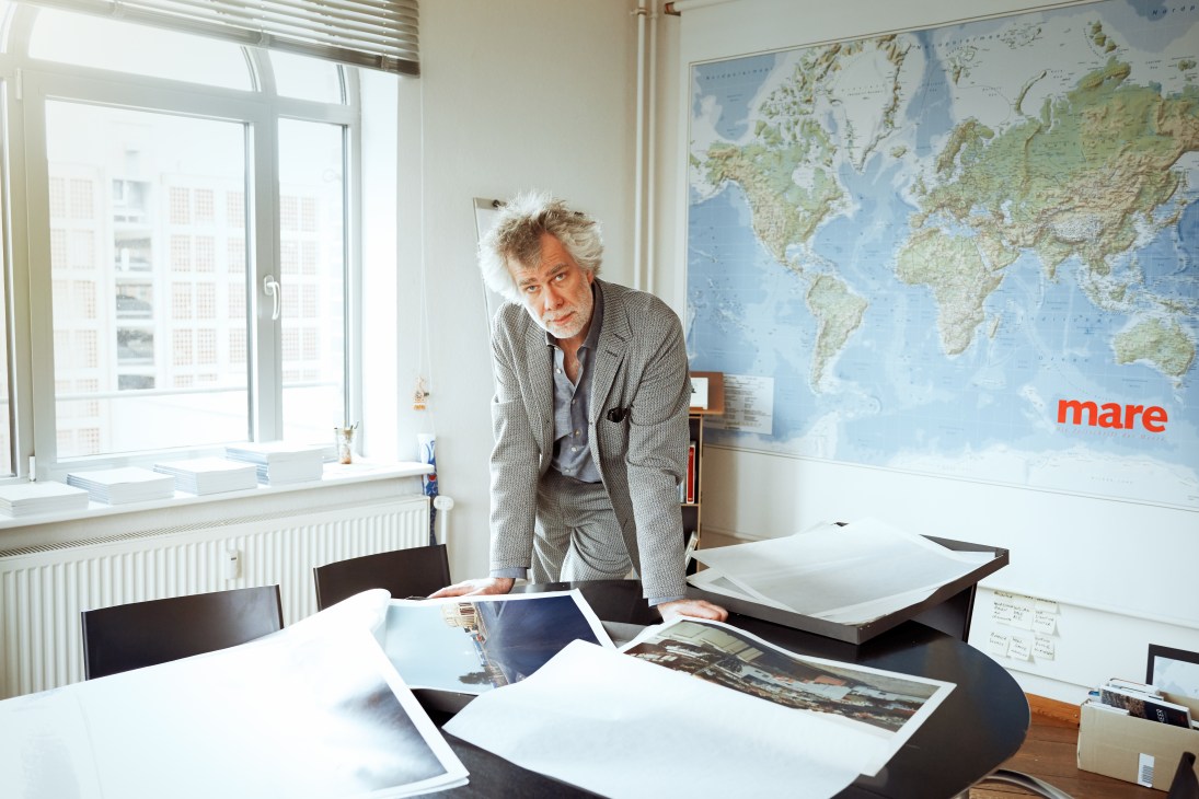 Nikolaus Gelpke, co-founder and editor in chief of Mare magazine, leans over a desk reviewing photographs with a world map visible on the wall behind him.